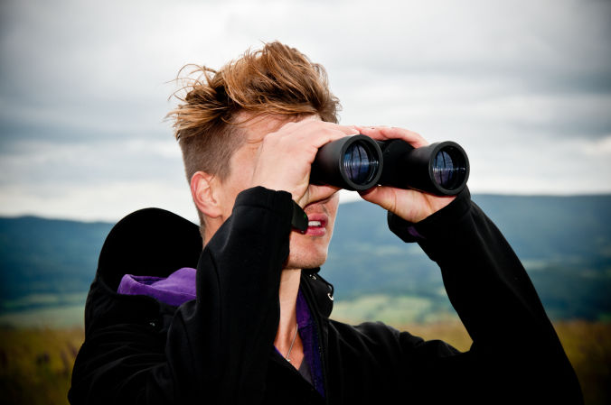 Mensch schaut durchs Fernglas, im Hintergrund eine grüne Hügellandschaft